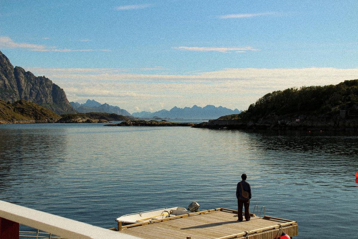 Person standing on a wooden pier facing a calm bay and mountains in Lofoten