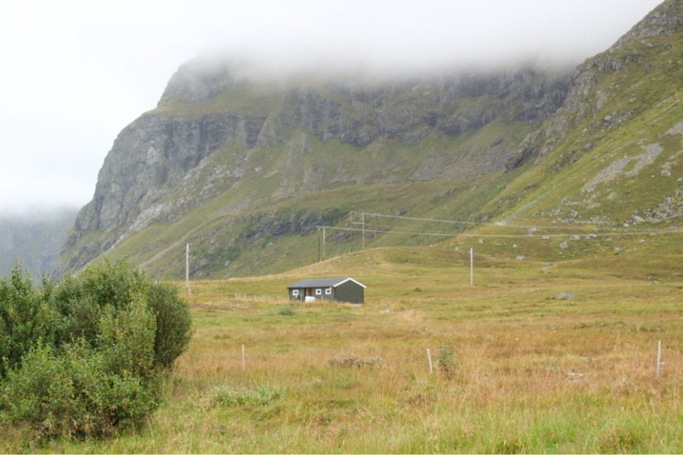 Small house on a green ridge below cloud-capped cliffs in Lofoten
